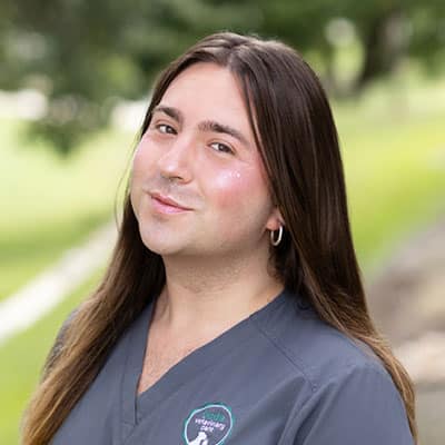 Person with long brown hair wearing gray scrubs and a hoop earring, standing outdoors with a blurred green background, smiling gently at the camera.