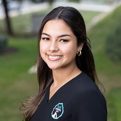 A young woman with long, dark hair and gold hoop earrings smiles outdoors. She is wearing a black top with a logo on it and standing in front of a blurred green background.