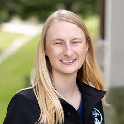 A person with long blonde hair smiles outdoors, wearing a black jacket with a circular logo, standing in front of a blurred green background.