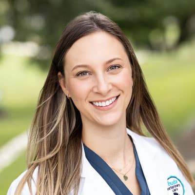 A woman with long brown hair wearing a white lab coat smiles outdoors, with green trees and grass blurred in the background.