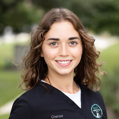 A young woman with wavy brown hair smiles outdoors, wearing a black medical scrub top with a name tag that says “Chenoa.” The background is blurred greenery.