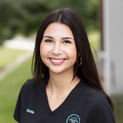 A woman with long dark hair, wearing hoop earrings and black scrubs, smiles outdoors. Her top has an embroidered logo and the name “Gabriela.” The background is blurred greenery and a building.