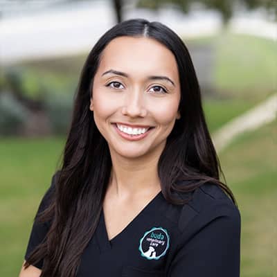 A woman with long dark hair, wearing a black scrub top with a "Buda Veterinary Clinic" logo, smiles at the camera while sitting outdoors with a green, blurred background.