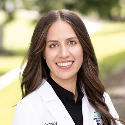 A woman with long brown hair smiles outdoors, wearing a white lab coat over a black shirt. The background is blurred greenery and a pathway.