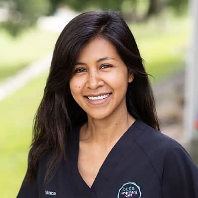 A woman with long dark hair, wearing black scrubs with a “buda veterinary care” logo, stands outdoors and smiles at the camera.