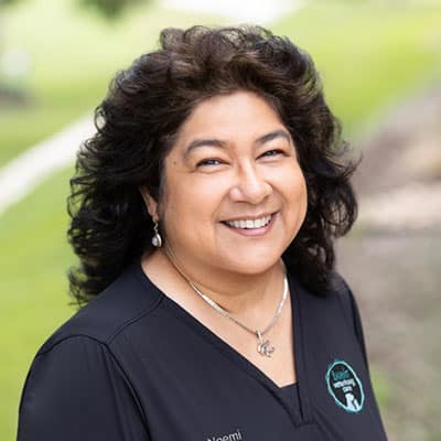 A smiling woman with curly dark hair wearing a black top and silver jewelry, standing outdoors with greenery in the background.