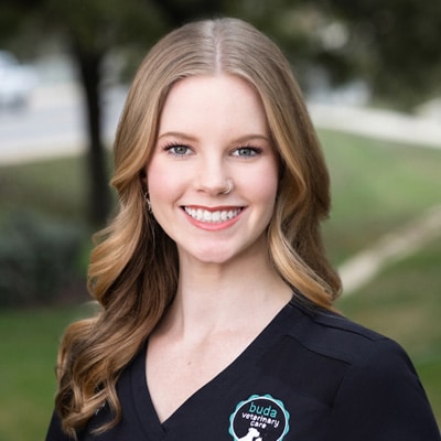 A smiling woman with long wavy blonde hair wearing a black veterinary uniform and a nose ring, standing outdoors with greenery in the background.