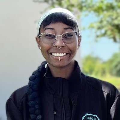 A person with glasses, facial piercings, and a long braided hairstyle smiles outdoors. They wear a light headband and a dark jacket with a circular logo on the chest. The background is blurred greenery.