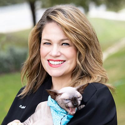 A smiling woman with wavy, light brown hair holds a hairless Sphynx cat wearing a blue bandana. The cat has its mouth open, baring its teeth. The background is outdoors with grass and trees.