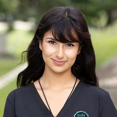 A young woman with long dark hair and a nose piercing smiles outdoors, wearing a black top with a small logo on it. The background is blurred green grass and trees.